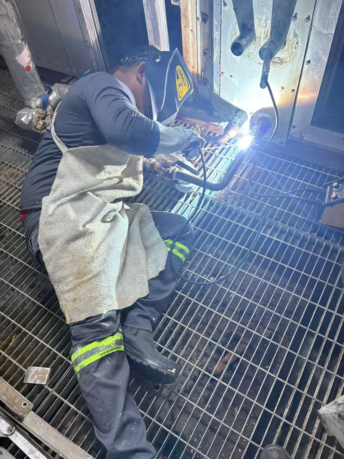 Profissional sentado sobre piso metálico, com máscara e avental, soldando conexões na parte inferior de um equipamento.