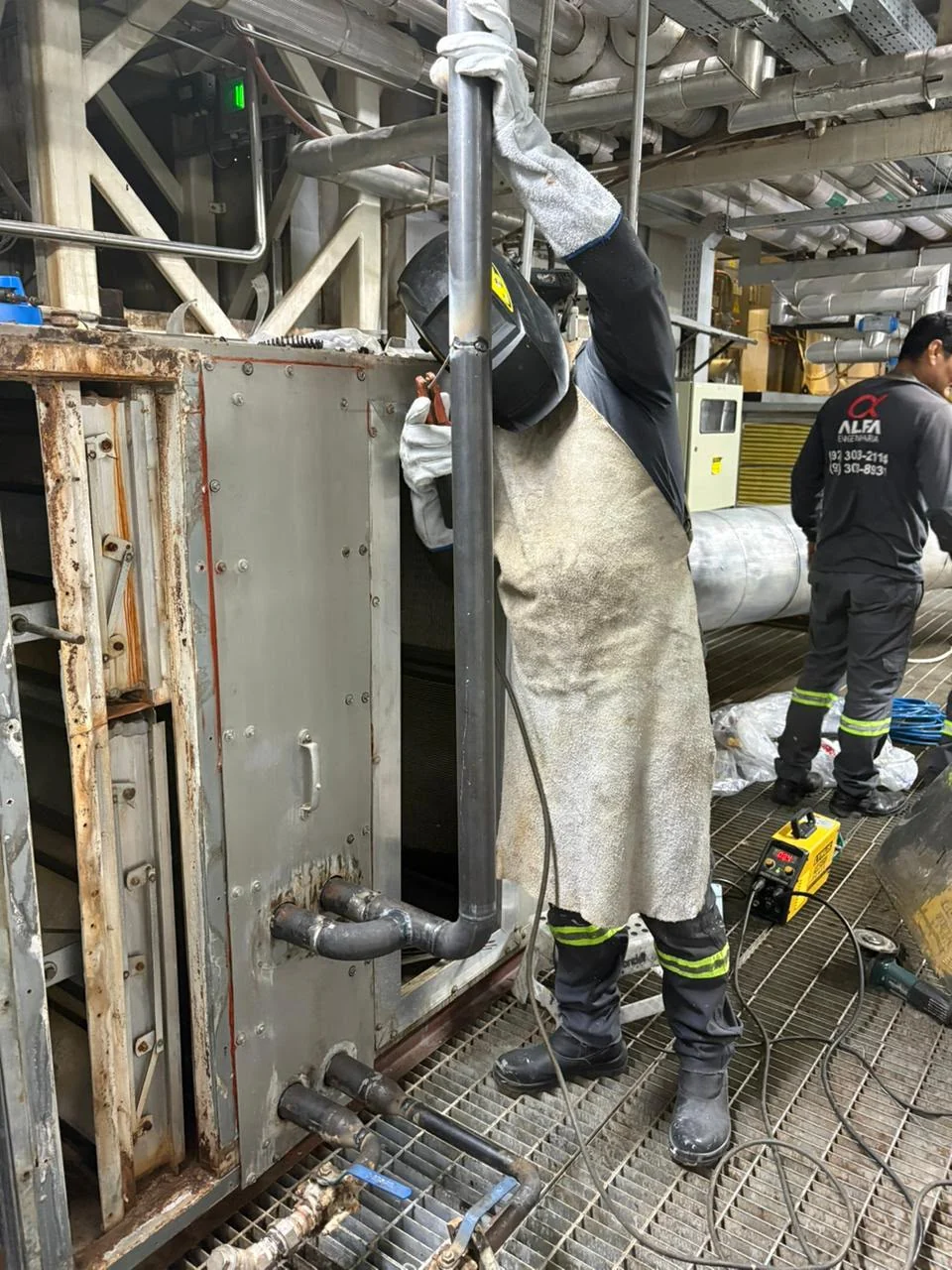 Profissional com máscara, luvas e avental trabalhando em uma tubulação vertical junto a um equipamento.