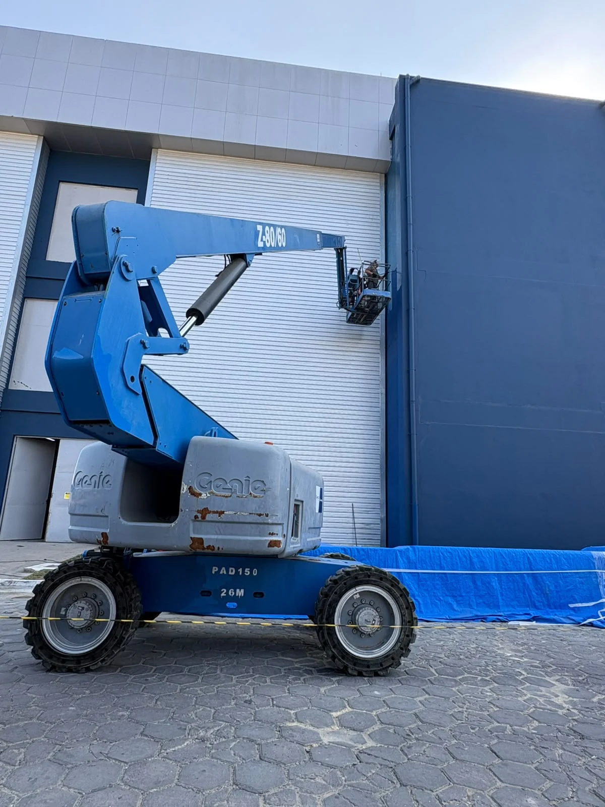 Plataforma elevatória junto a uma fachada azul e branca, com lona azul protegendo a base da parede.