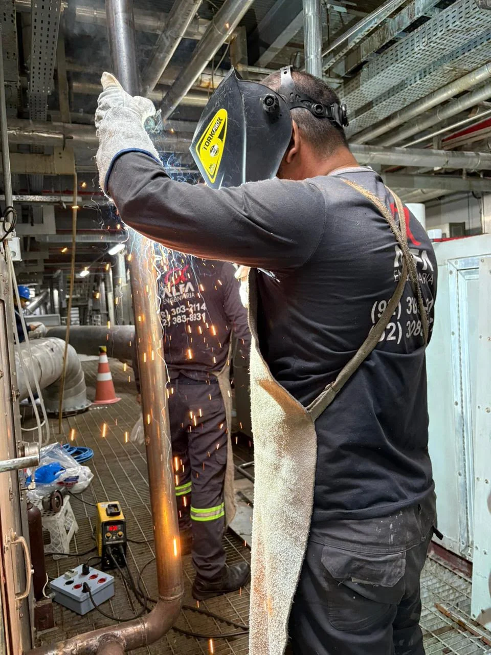 Profissional da Alfa visto de costas, com máscara e luvas, executando soldagem em uma tubulação vertical.