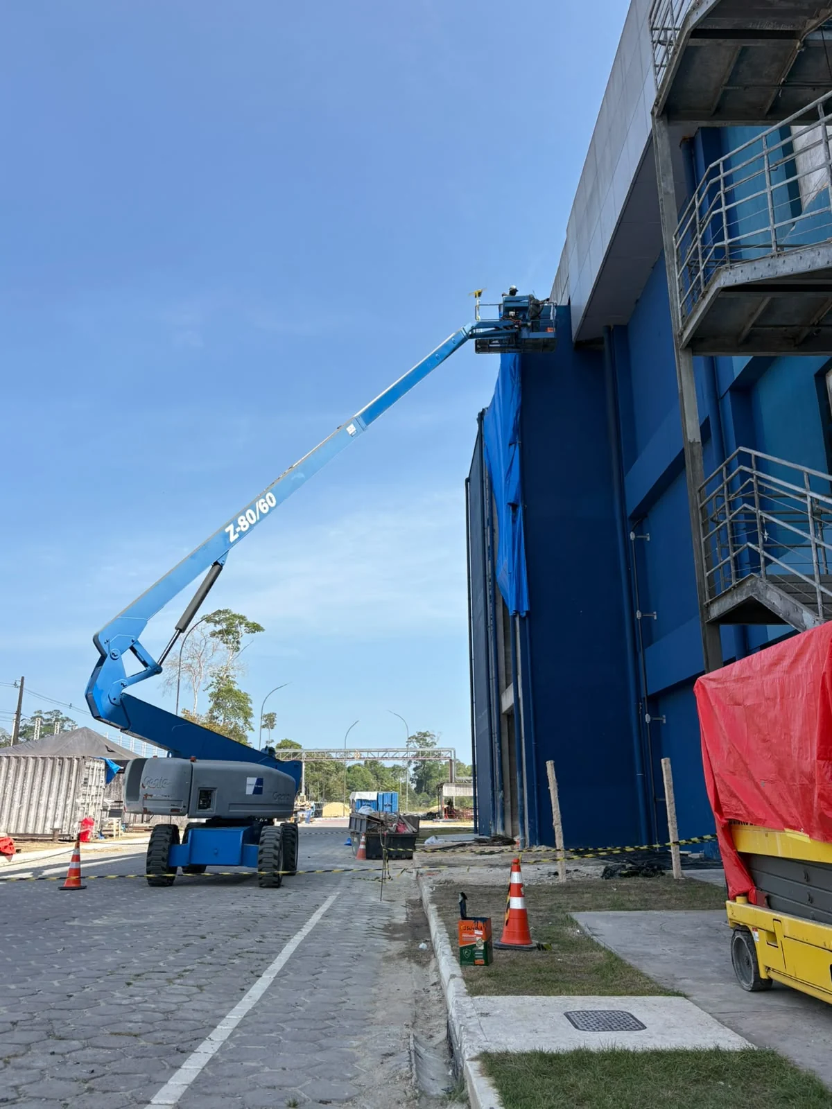 Plataforma elevatória com o braço estendido até a parte superior de uma fachada azul, em uma área delimitada por cones.